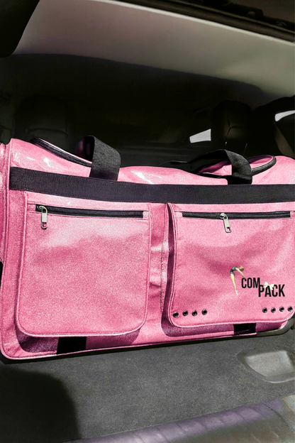 Pink ComPack rolling dance competition bag in car trunk, packed with dance costumes and gear, featuring glittery PU-coated Oxford fabric, wheels, folded garment rack, and carry handles.